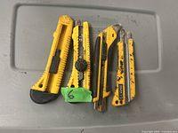 Five yellow box cutters laid out horizontally on a gray plastic surface, showing varying levels of wear and used blade adjustment mechanisms.