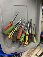 Photo showing lot of 11 used screwdrivers with various handle colors and blade types on a gray surface.