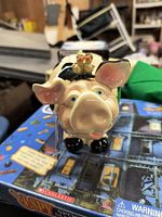 Front view of cream-colored piggy bank with blue eyes and two birds figurine on head, showing detailed facial features and black hooves.