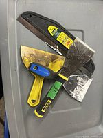 Photo showing three used scrapers on a gray surface with visible paint marks and wear on blades.