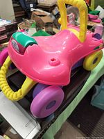 Pink riding toy with yellow handlebars, purple and green accents, and yellow wheels shown from side angle.