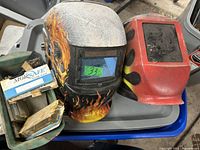Two welding helmets: one with flame design and blue lens, the other plain red with worn lens. Small container filled with assorted welding glass plates.
