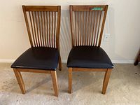 Two wood dining chairs with black faux leather seats shown from the front on carpeted floor.