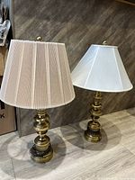 Two table lamps with matching brass bases and different shades (pleated beige and white fabric) on a wooden floor against wood panel background.