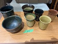 Seven ceramic items displayed on wooden table: one bowl, three vases, three cups.