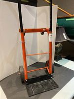 Front view of the convertible hand cart showing orange frame, black handles, and black base plate with two large wheels.