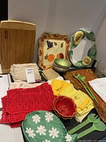 Photo showing wooden cutting boards, decorative plates with fruit and sunflower designs, colorful ceramic bowls, assorted table linens including red crocheted cloth and yellow embroidered napkins, and green plastic salad serving spoons.