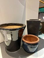Photo showing all three plant pots together on a shelf: large glossy black pot, tall matte black pot, and small navy pot with terracotta rim.
