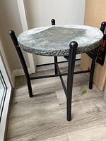 Full view of the round concrete top table with black metal legs on wood flooring next to window.