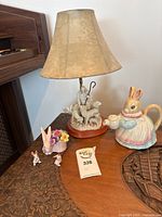 Photo showing the bunny teapot, lamp with shepherd and lamb figurines, and three small bunny porcelain figurines on wooden furniture.