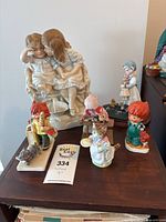 Photo showing a variety of vintage porcelain and Hummel figurines arranged on a wooden table, including a girl whispering to a boy, Hummel children with baskets and animals