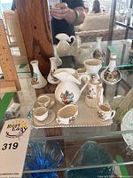 Photo showing various miniature ceramic teacups, pitchers, and small vases arranged on a glass shelf. Pieces are white ceramic with colorful coat of arms crests painted on them. Tray holding several small teacups and a pitcher is visible.