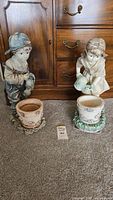 Two ornamental planters depicting a boy and a girl holding watering cans, standing against a wooden dresser on carpeted floor.