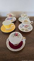 Five assorted floral tea cup and saucer sets arranged on wooden surface showing various colors and gold trim