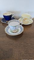 Three assorted vintage teacups and their matching saucers on a wooden surface showing different floral and gold rim designs.