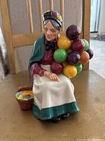 Front view of the Royal Doulton The Old Balloon Seller figurine showing the woman holding colorful balloons, seated wearing a green shawl and bonnet, with a basket of balloons nearby.