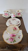 Photo of multiple Royal Albert teacups and saucers with pink rose floral patterns and gold trim arranged on a wooden surface