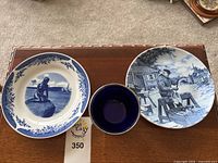 Two large decorative plates and one small bowl displayed on a table, showing detailed blue and white art depicting Danish scenes and motifs.