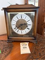 Front view of square wooden clock with glass door closed, showing clock face with Roman numerals and intricately decorated corners.