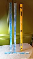 Two blue Empire drywall T-squares and one yellow Stanley level standing upright against a green wall on a blue cloth.
