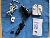 Photo showing two sets of Apple wired earphones (one in white plastic case, one loose), one set of black generic wired earphones, and one black USB car charger on blue cloth background.