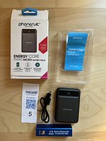 PhoneSuit Energy Core Studio Micro Battery Pack and accessories laid out with box, user manual, USB cable, and Mazda branded smaller battery pack on wooden surface.