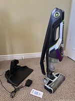 Photo showing the handheld vacuum cleaner standing upright next to the charger and power adapter on carpet.
