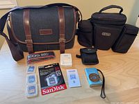 Full lot view showing camera bags, camera strap, memory cards in plastic cases, sealed SanDisk 64GB card, Canon battery charger, and blue Canon compact camera.