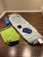 Camping gear set on wood floor: grey Traynor mummy sleeping bag laid open, blue sleeping mat rolled, bright green waterproof Baja bag rolled closed, small dark blue bowl and cup, blue MEC toiletry bag.