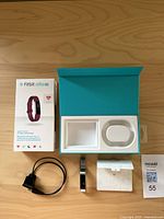 Fitbit Alta HR fitness tracker with silver metal mesh band, original box (empty insert), charging cable, instruction manual, and lot tag on wooden surface.