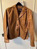 Front view of tan leather jacket hanging on black hanger, displaying button front closure and two front pockets.