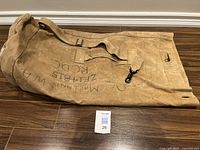 Brown canvas military duffel bag laid flat showing black writing and hook closure on wood floor.