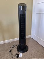 Front view of tall black tower fan with vertical vent and power cord deployed on carpeted floor.