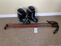 Pair of dark blue Dalbello ski boots and red youth ski poles arranged on beige carpet, showing overall appearance and size.