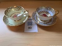 Two sets of fine china teacups and saucers displayed side by side on a wooden surface, showing detailed gold and floral patterns, one set cream colored with ornate gold scrollwork, the other white with gold floral border and pink rose center.