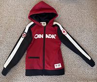 Front view of the Team Canada 2010 Olympic girls' jacket showing the hood, zipper, and patches on the sleeves.