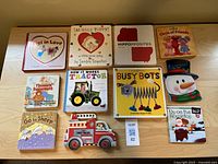 Top view of all thirteen toddler and preschool board books arranged in a rectangular pattern on a wooden surface. Visible titles include 'Pigs in Love,' 'Snuggle Puppy!,' 'Hippopposites,' 'Circle of Friends,' 'Thomas' Snowsuit,' 'How It Works Tractor,' 'Busy Bots,' 'Snowman shaped book,' 'Go to Sheep,' 'Fire Truck shaped book,' and 'Up on the Housetop.'