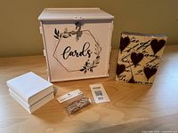 Photo of white metal wedding card box, ceramic heart decoration, miniature clothespins, and small envelopes arranged on a wooden surface.