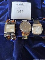 Five fashion watches arranged on a dark blue fabric surface. Visible watches show variations in band material and dial face design: tortoise shell plastic, metal bands, scripted logo dial, crystal accents.