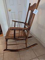 Side profile of the antique wooden rocking chair highlighting the caned seat, spindled armrest supports, and curved wooden rockers.