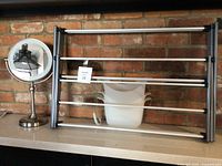 Battery operated magnifying mirror, 3-section white plastic shower caddy with metal frame, and foldable laundry rack shown on a shelf against a brick wall