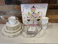 Full set of dishes showing 4 dinner plates, 4 dessert plates, 4 cups and saucers arranged with glass cake plate and cupcake picture in the background