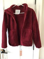 Front view of the burgundy fleece sweater hanging on a door. Shows soft texture, front zipper, and pockets.