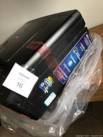 Side angle view of black Brother printer wrapped in plastic with taped sections to keep parts closed during transport.