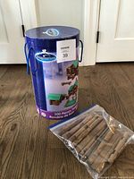 Full view of the tall cylindrical container packaging labeled Imagitarium with handle and image of a timber log structure made from the set, along with clear plastic bag containing wooden log pieces on top of the cylinder.