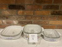 Three Corningware casserole dishes of different sizes with lids decorated with blue cornflower pattern sitting on a white surface against a brick wall background.