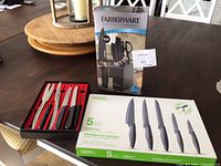 Three boxed sets of kitchen cutting utensils shown on a table including the steak utensil set in an open black and red case, the Everyday Essentials knife set in open green and white box, and the Farberware knife set in gray box.