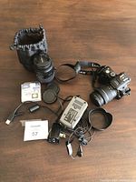 Overview of camera body, two lenses, filter, charger, lens hood, battery and headphones on table with lot tag