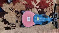 Top angled view of the Plasma Car ride-on toy showing blue body, pink seat, black steering wheel and front wheels on patterned carpet.