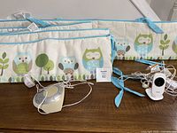 Photo of two owl pattern crib bumper pads with blue trim, a white baby monitor, and a circular sound machine on a wooden surface